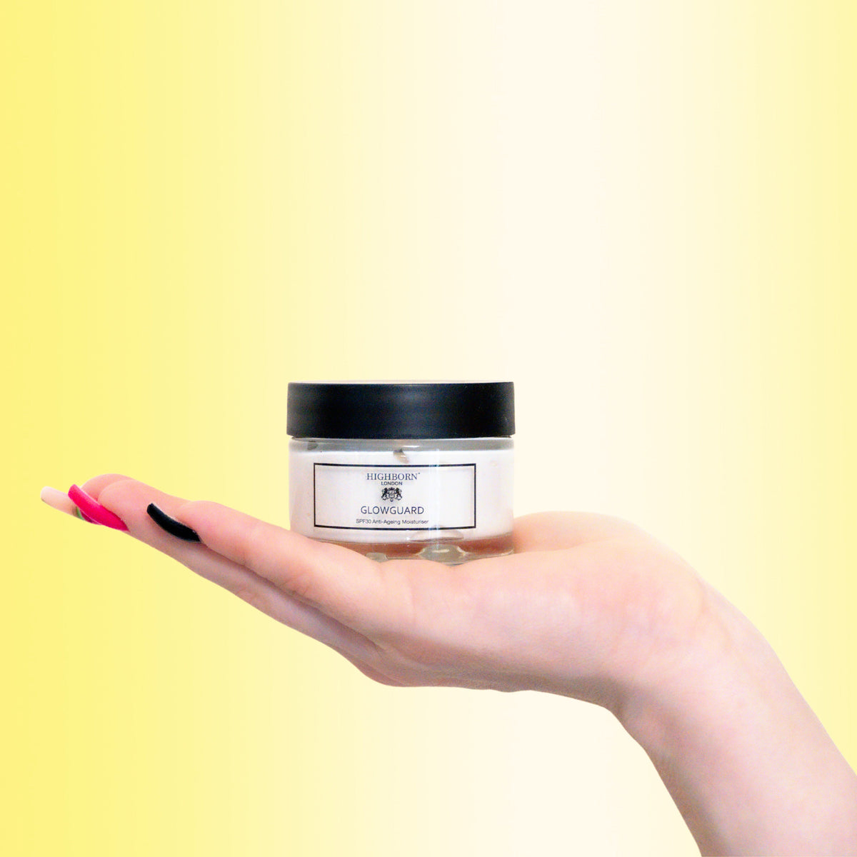Open hand holding a single Highborn London GLOWGUARD SPF30 Anti-Ageing Moisturiser jar against a gradient yellow studio background